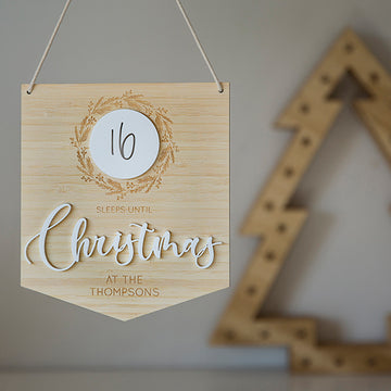 Personalised Christmas Countdown Banner: Wooden sign with "16 sleeps until Christmas at the Thompsons" in white text, decorated with a wreath and hung on string. A blurred wooden Christmas tree appears in the background.