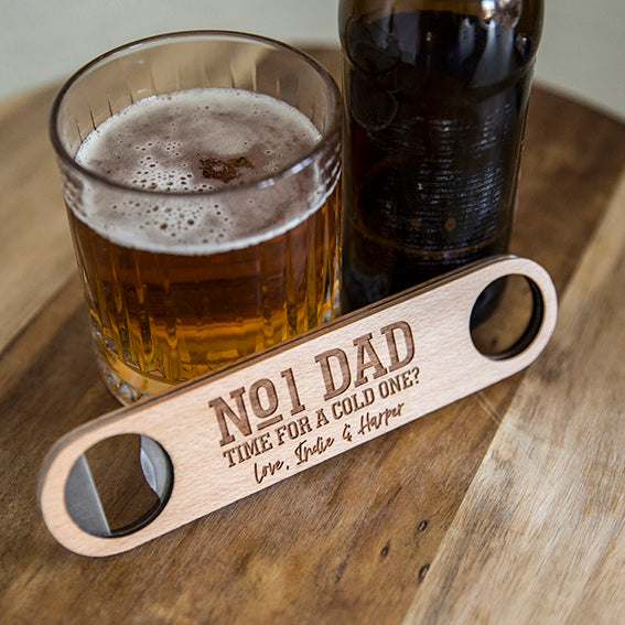 A dark beer bottle, a glass of beer, and a Personalised Bottle Opener (3 designs available) engraved with "No1 Dad Time for a cold one? Love, India & Harper" are set on a wooden table—an ideal gift for men who value thoughtful details.