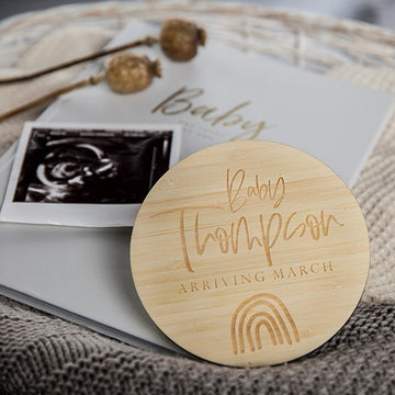 The Personalised Rainbow Announcement Card, engraved with "Baby Thompson Arriving March" and a rainbow, sits beside a sonogram and "Baby" card on a textured surface with dried flowers—perfect for your rainbow baby announcement.