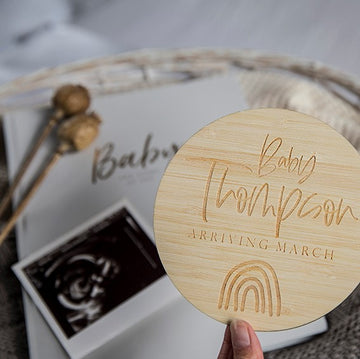 A hand holds a Personalised Rainbow Announcement Card engraved with "Baby Thompson Arriving March" and a rainbow, ideal for a rainbow baby announcement. In the background are a baby book, ultrasound photo, and dried poppy pods.
