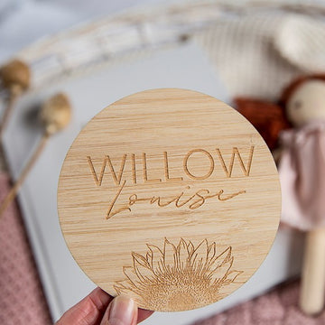 A hand displays the Personalised Sunflower Plaque, engraved with WILLOW Louise above a sunflower design. With a doll, book, and dried plants softly blurred behind, this charming plaque is the perfect birth announcement.