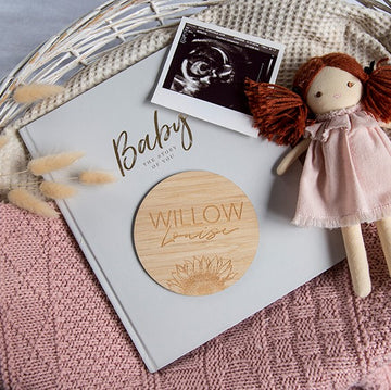 A baby memory book, a Personalised Sunflower Plaque engraved with Willow Louise, a baby ultrasound photo, and a doll with red yarn hair and pink dress rest on a pink knitted blanket in a wicker basket.