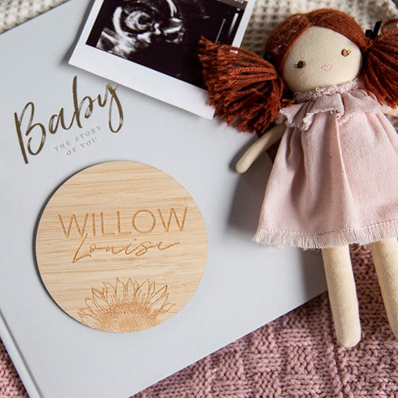 A cloth doll in a pink dress rests beside a baby book, a Personalised Sunflower Plaque engraved with Willow Louise, and a black-and-white ultrasound photo—ideal for a charming birth announcement on a pink knitted blanket.