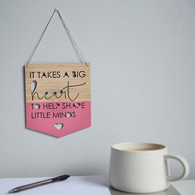 A bamboo banner saying It takes a big heart to help shape little minds, with a pink heart cut out, half painted in pink. Hanging above a teacher's desk.
