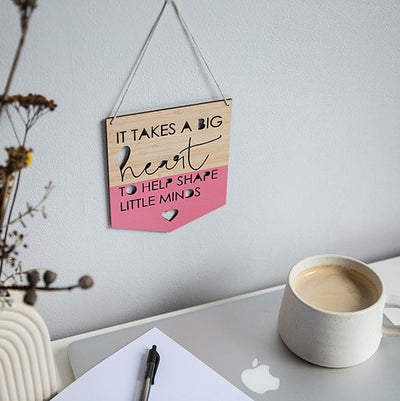 A bamboo banner saying It takes a big heart to help shape little minds, with a pink heart cut out, half painted in pink. Hanging above a teacher's desk.