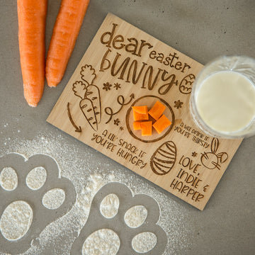 The Personalised Easter Bunny Treat Board, featuring a sweet message and etched design, is perfect for creating a festive Easter scene with milk, carrot cubes, whole carrots, and flour paw prints on the counter.
