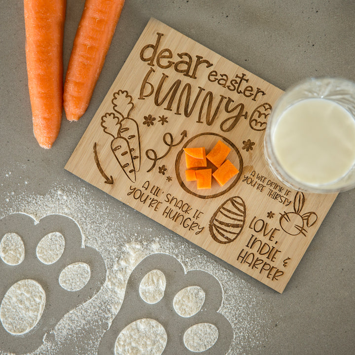 The Personalised Easter Bunny Treat Board, featuring a sweet message and etched design, is perfect for creating a festive Easter scene with milk, carrot cubes, whole carrots, and flour paw prints on the counter.