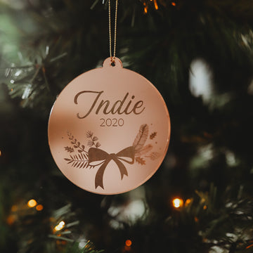 The Bow Christmas Bauble is a round, gold ornament engraved with "Indie 2020," featuring floral designs and a bow. It hangs on a lit Christmas tree with green pine needles—a perfect keepsake, complete with gift pouch.
