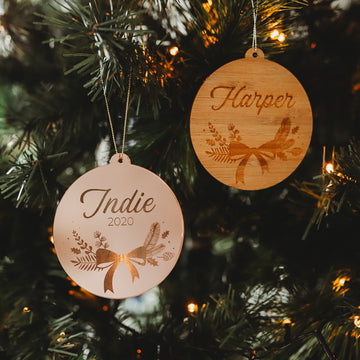 Two Bow Christmas Bauble ornaments hang on a tree, engraved with “Indie 2020” and “Harper,” featuring botanical designs, bows, and glowing lights. Each comes in a keepsake gift pouch.
