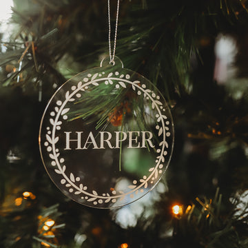 A Personalised Frosted Christmas Bauble with a white leafy border and the name HARPER hangs among glowing lights and pine needles.