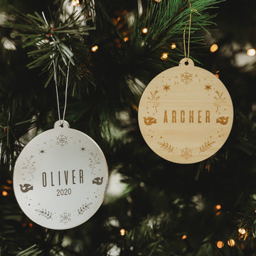 Two round ornaments hang on the Christmas tree—a white personalised wreath reading OLIVER 2020, and a Winter Wonderland Christmas Bauble marked ARCHER. Both glow amid twinkling lights and festive patterns.