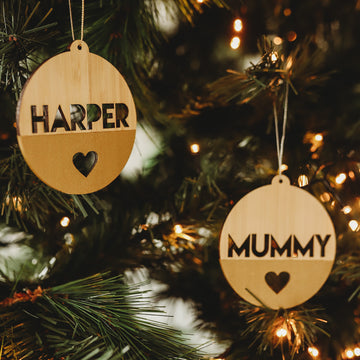 Two Original Personalised Christmas Baubles hang on the tree, one reading HARPER and the other MUMMY, each with a heart cutout—adding a special family touch to your Christmas decorations.