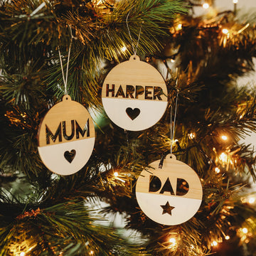 Three The Original Personalised Christmas Baubles hang on a Christmas tree, each round and wooden with cut-out words: “MUM” with a heart, “HARPER” with a heart, and “DAD” with a star. Warm white lights illuminate the tree.