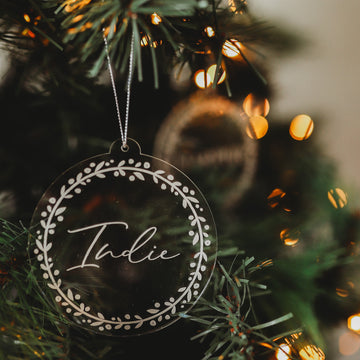 The Personalised Frosted Christmas Bauble features a round, clear acrylic design engraved with the name Indie in cursive and a white leafy border, hanging on a Christmas tree amid soft lights.