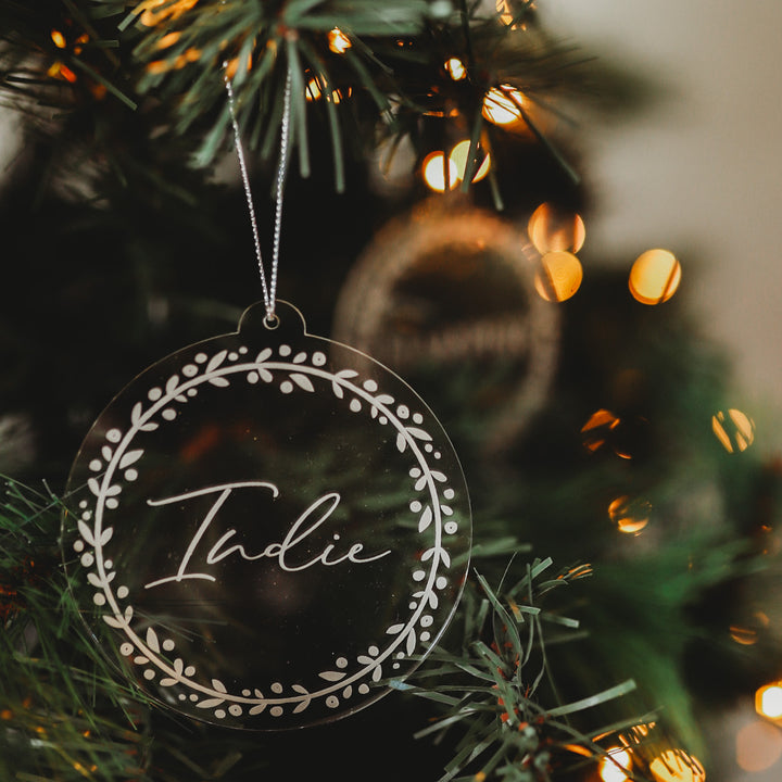 The Personalised Frosted Christmas Bauble features a round, clear acrylic design engraved with the name Indie in cursive and a white leafy border, hanging on a Christmas tree amid soft lights.