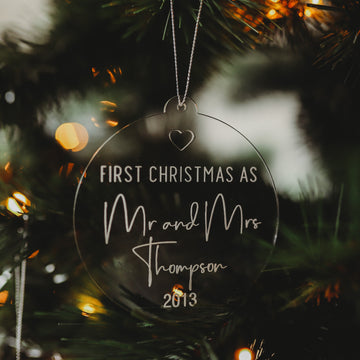 The Engaged/Mr & Mrs Christmas Bauble hangs on a tree, engraved with “First Christmas as Mr and Mrs Thompson 2013,” surrounded by warm lights and pine needles, making a meaningful keepsake gift.