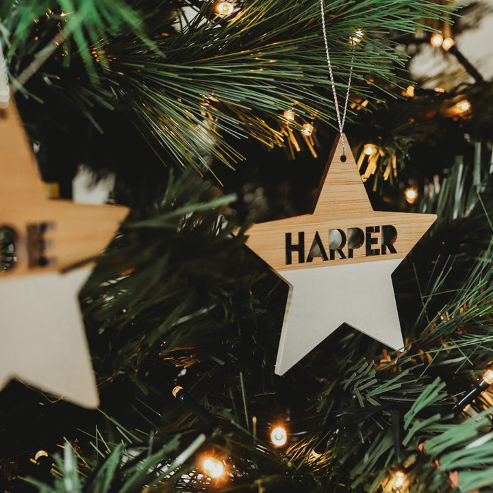 A Personalised Christmas Stars ornament featuring the name Harper hangs on a tree with green branches and glowing string lights, adding charm to your festive décor.