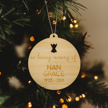 The "In Loving Memory Christmas Bauble" is a round wooden ornament displayed on a tree, featuring the inscription "In loving memory of Nan Grace 1925-2011" with an angel silhouette above and twinkling lights in the background.