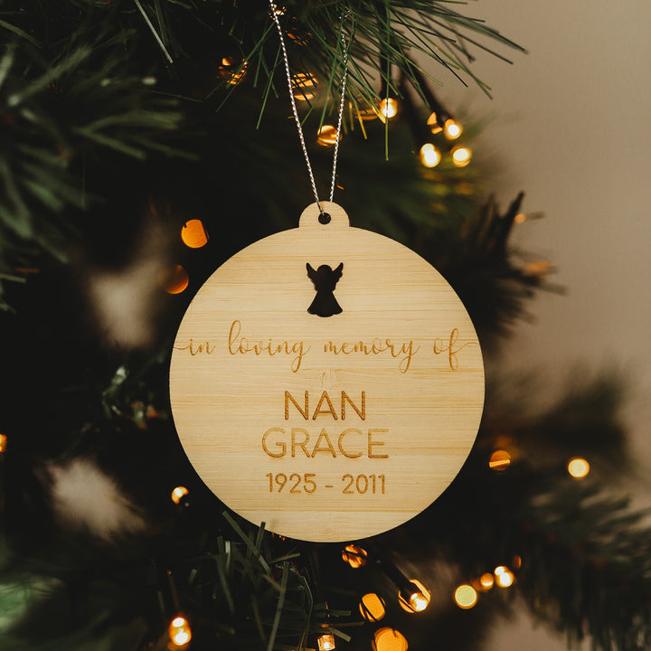 The "In Loving Memory Christmas Bauble" is a round wooden ornament displayed on a tree, featuring the inscription "In loving memory of Nan Grace 1925-2011" with an angel silhouette above and twinkling lights in the background.