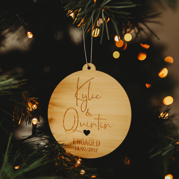 An Engaged/Mr & Mrs Christmas Bauble hangs on a tree, engraved with "Kylie & Quentin, Engaged 14/07/2012" and heart designs—a perfect keepsake gift illuminated by warm fairy lights.