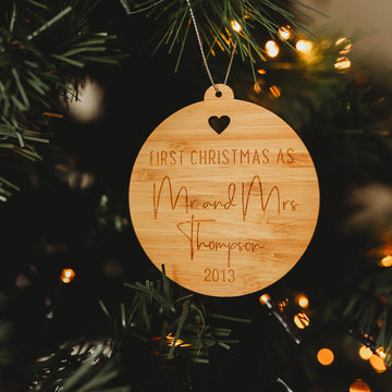 The Engaged/Mr & Mrs Christmas Bauble, a round wooden ornament with a heart cutout, hangs on a tree surrounded by green pine needles and warm lights. Engraved with First Christmas as Mr and Mrs Thompson 2013, it&