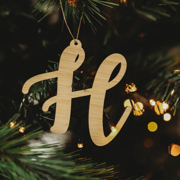 A Letter Christmas Ornament hangs from a tree branch, nestled among pine needles and glowing fairy lights—one of many charming ornaments ideal for personalizing your holiday décor.