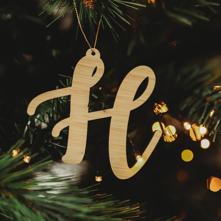 A Letter Christmas Ornament hangs from a tree branch, nestled among pine needles and glowing fairy lights—one of many charming ornaments ideal for personalizing your holiday décor.