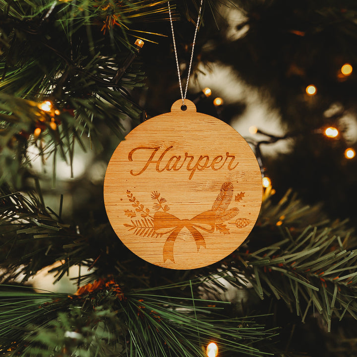 The Bow Christmas Bauble, a round bamboo ornament engraved with "Harper" and topped with a festive bow and pine branches, hangs from a tree with warm white lights—perfectly giftable in its keepsake pouch.