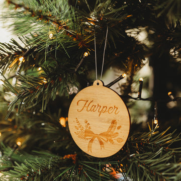 The Bow Christmas Bauble, a round bamboo ornament engraved with botanical designs and the name Harper, hangs on a tree adorned with white lights, making a charming keepsake gift pouch addition.