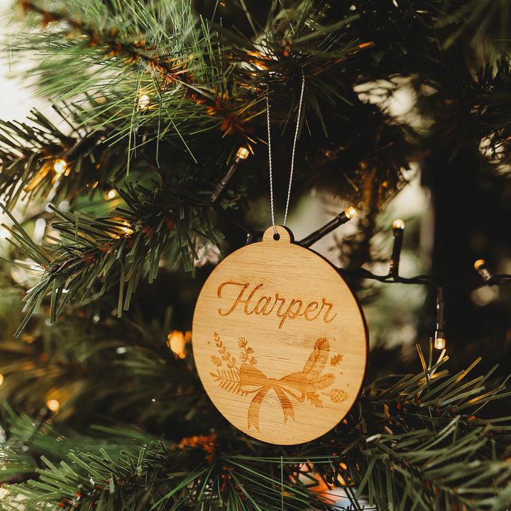 The Bow Christmas Bauble, a round bamboo ornament engraved with botanical designs and the name Harper, hangs on a tree adorned with white lights, making a charming keepsake gift pouch addition.