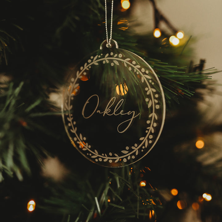 A Personalised Frosted Christmas Bauble, engraved with the name Oakley and a leafy border, hangs on a tree branch illuminated by warm white lights.