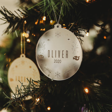 The Winter Wonderland Christmas Bauble hangs on a tree adorned with lights, beside a personalised wreath ornament. Another blurred bauble appears in the background, adding to the festive holiday scene.