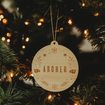 The Winter Wonderland Christmas Bauble, engraved with the name ARCHER and surrounded by stars, leaves, and two birds, hangs on a glowing Christmas tree.