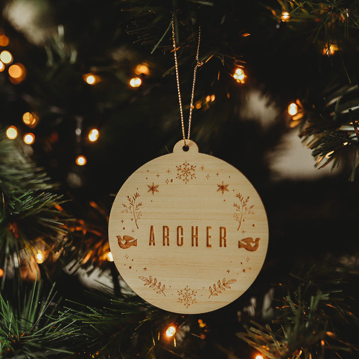 The Winter Wonderland Christmas Bauble, engraved with the name ARCHER and surrounded by stars, leaves, and two birds, hangs on a glowing Christmas tree.