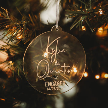 The Engaged/Mr & Mrs Christmas Bauble hangs on a Christmas tree with “Kylie & Quintin ENGAGED 14/07/2023” and a heart engraved, making a beautiful keepsake gift among green branches and warm white lights.