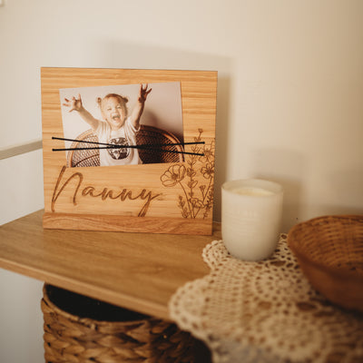 A dark bamboo photo board with Nanny and a floral piece engraved. A photo of a girl is on it, held with black elastic. Sitting on a shelf. 