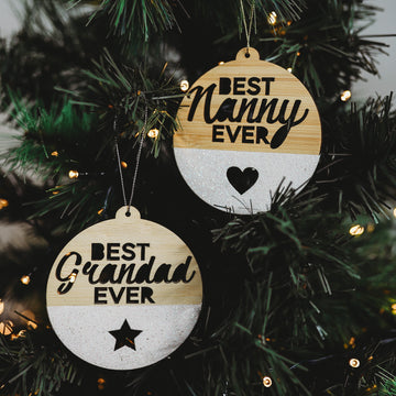 Two Best Ever Christmas Bauble ornaments hang on a tree—each reads "Best Nanny Ever" or "Best Grandad Ever," features heart and star cutouts, and makes a unique, glittery personalised gift for loved ones.