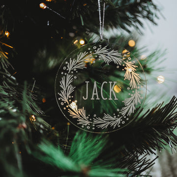 The Pine Frosted Christmas Bauble, etched with the name JACK and framed by a white leafy wreath design, hangs on a green branch and glows in the light—making it a perfect personalised ornament for your tree.