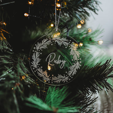 The Pine Frosted Christmas Bauble, etched with Ruby and a leafy wreath design, hangs on a tree adorned with glowing fairy lights.