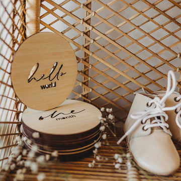 A round light bamboo set of baby milestone markers sitting on a shelf with a floral arrangement and some baby shoes. Visible signs say hello world and one month. 