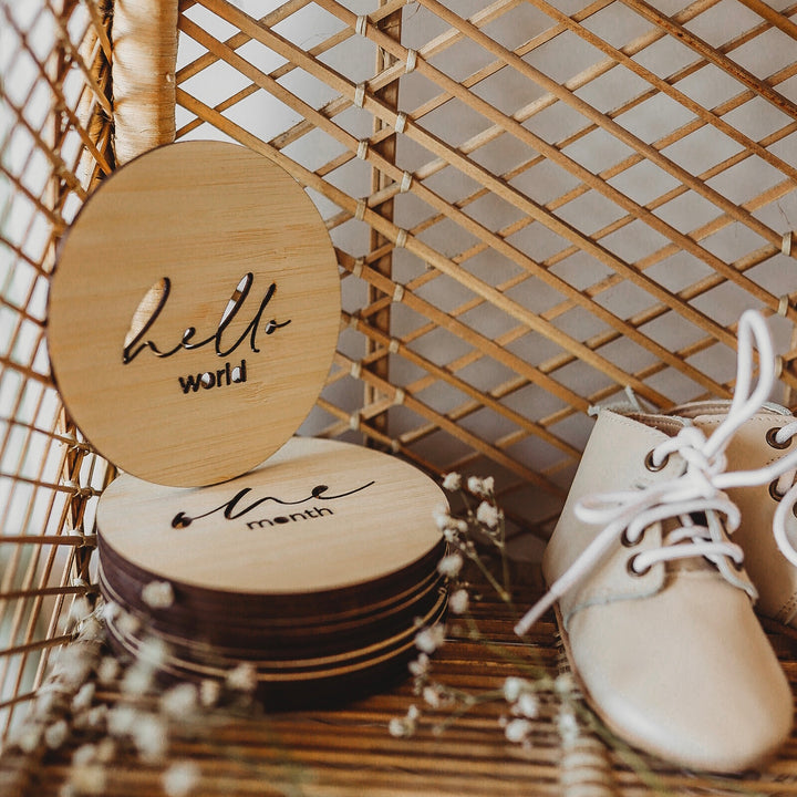 A round light bamboo set of baby milestone markers sitting on a shelf with a floral arrangement and some baby shoes. Visible signs say hello world and one month. 