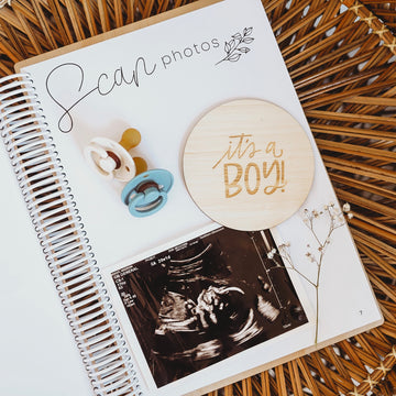 A spiral-bound scrapbook from the Lovely Lettering Pregnancy Milestones Set is open to a page with an ultrasound photo, wooden "It’s a BOY!" milestone cards, two pacifiers, and baby&