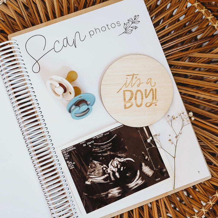 A spiral-bound scrapbook from the Lovely Lettering Pregnancy Milestones Set is open to a page with an ultrasound photo, wooden "It’s a BOY!" milestone cards, two pacifiers, and baby&