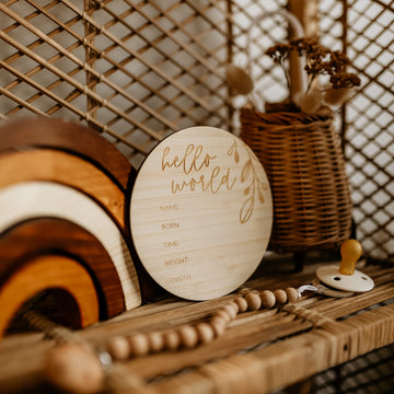 The Round Birth Announcement Card, crafted from eco-friendly bamboo, is displayed on a wicker shelf with wooden rainbows, a beaded pacifier clip, and a basket of dried flowers.