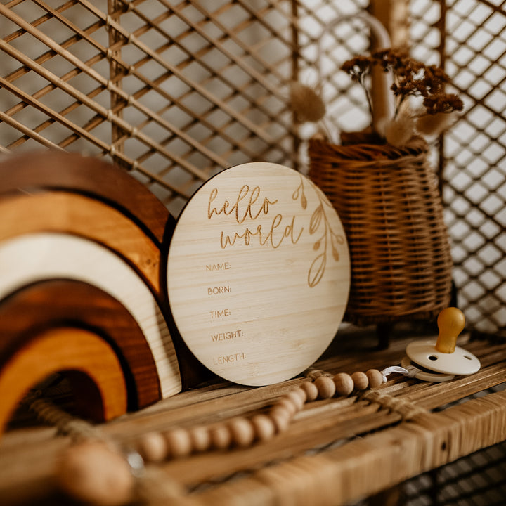 The Round Birth Announcement Card, crafted from eco-friendly bamboo, is displayed on a wicker shelf with wooden rainbows, a beaded pacifier clip, and a basket of dried flowers.