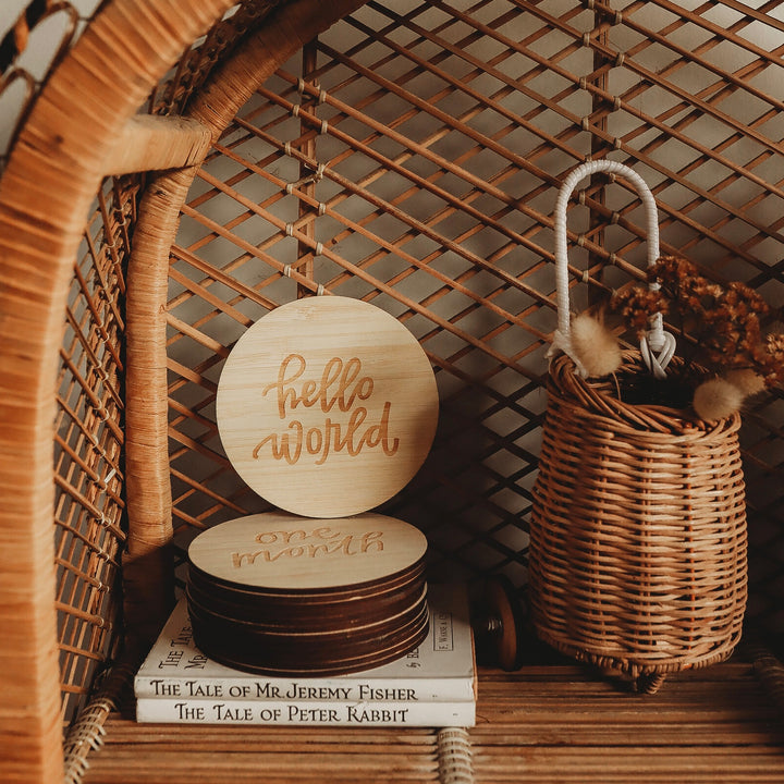 A woven rattan shelf displays Lovely Lettering Baby Milestone Sets wooden cards reading “hello world” and “one month,” plus a basket with dried flowers and two books: The Tale of Mr. Jeremy Fisher and The Tale of Peter Rabbit.