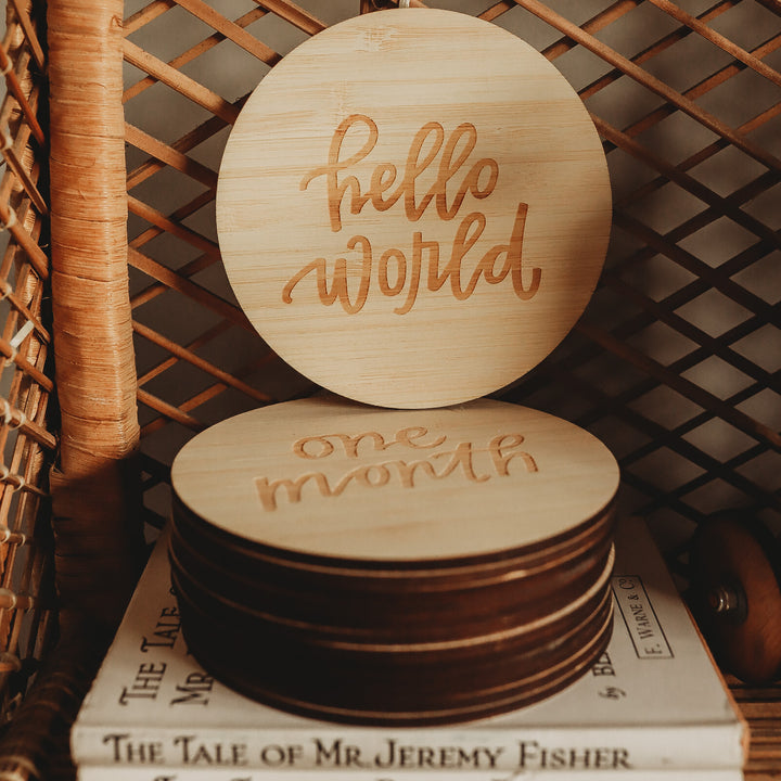 The Lovely Lettering Baby Milestone Sets feature round wooden cards with engraved text, stacked on books. The top card says "hello world," the second "one month," and a woven wicker chair is visible in the background.