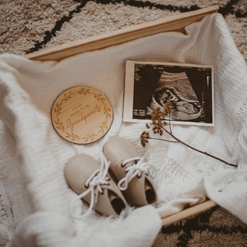 A wooden box lined with white cloth holds a baby ultrasound photo, tan baby shoes with white laces, a small dried flower, and a Personalised Grandparent/Godparent Card announcing “You’re going to be grandparents.”.