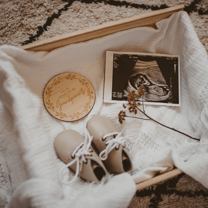 A wooden box lined with white cloth holds a baby ultrasound photo, tan baby shoes with white laces, a small dried flower, and a Personalised Grandparent/Godparent Card announcing “You’re going to be grandparents.”.