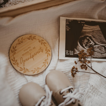 A Personalised Grandparent/Godparent Card made from bamboo is engraved with “You’re going to be grandparents” and displayed with a baby ultrasound photo, dried flowers, and baby shoes on soft cream-coloured fabric.